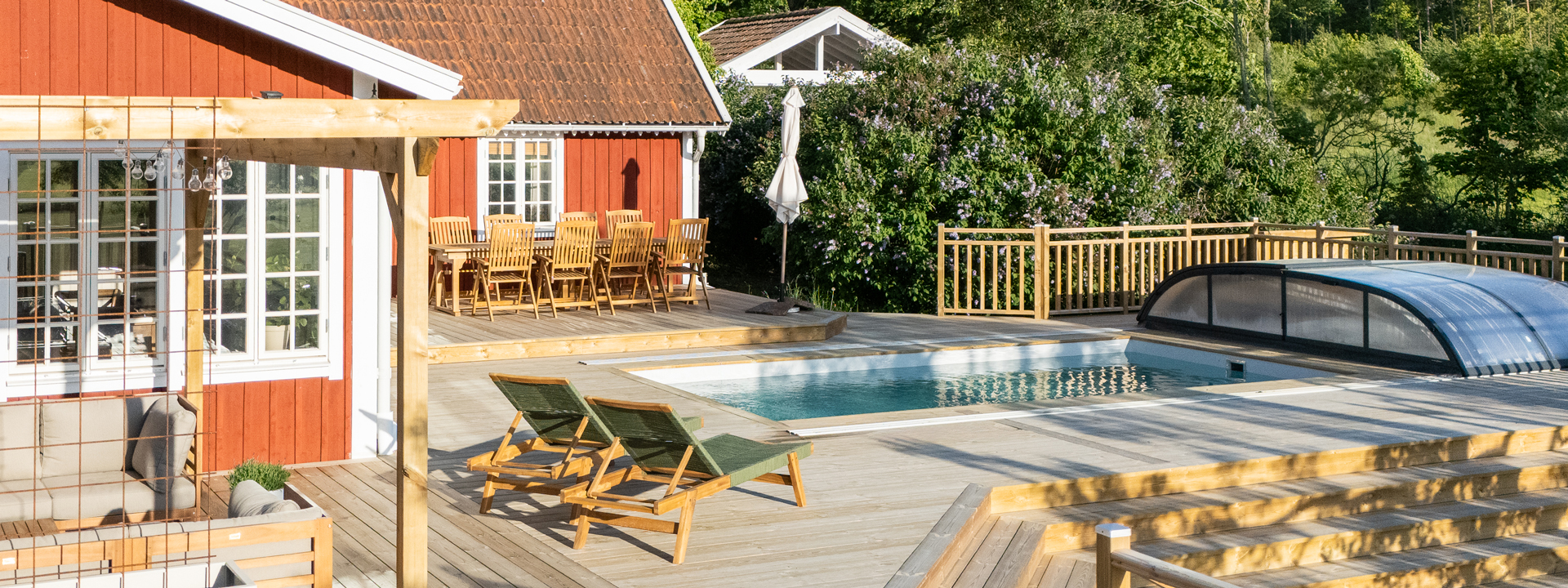 Ett trött hus med en stor altan i trä, och en pool, samt utemöbler och skog