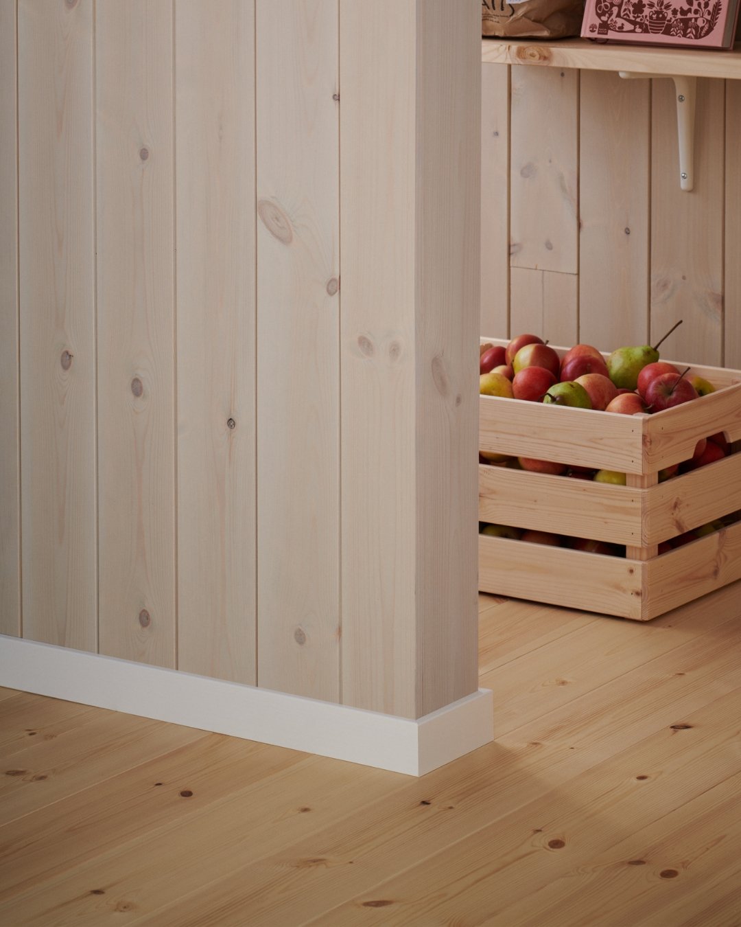 Vägg med panel, trägolv och vita lister, samt en trälåda med äpplen