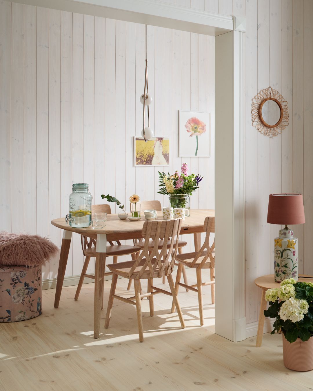 Matplats med vita träpanelen Sjöbod Snö på väggarna, runt träbord och matchande stolar. På bordet står blommor och en glasbehållare med vatten, och dagsljus faller in över det ljusa trägolvet