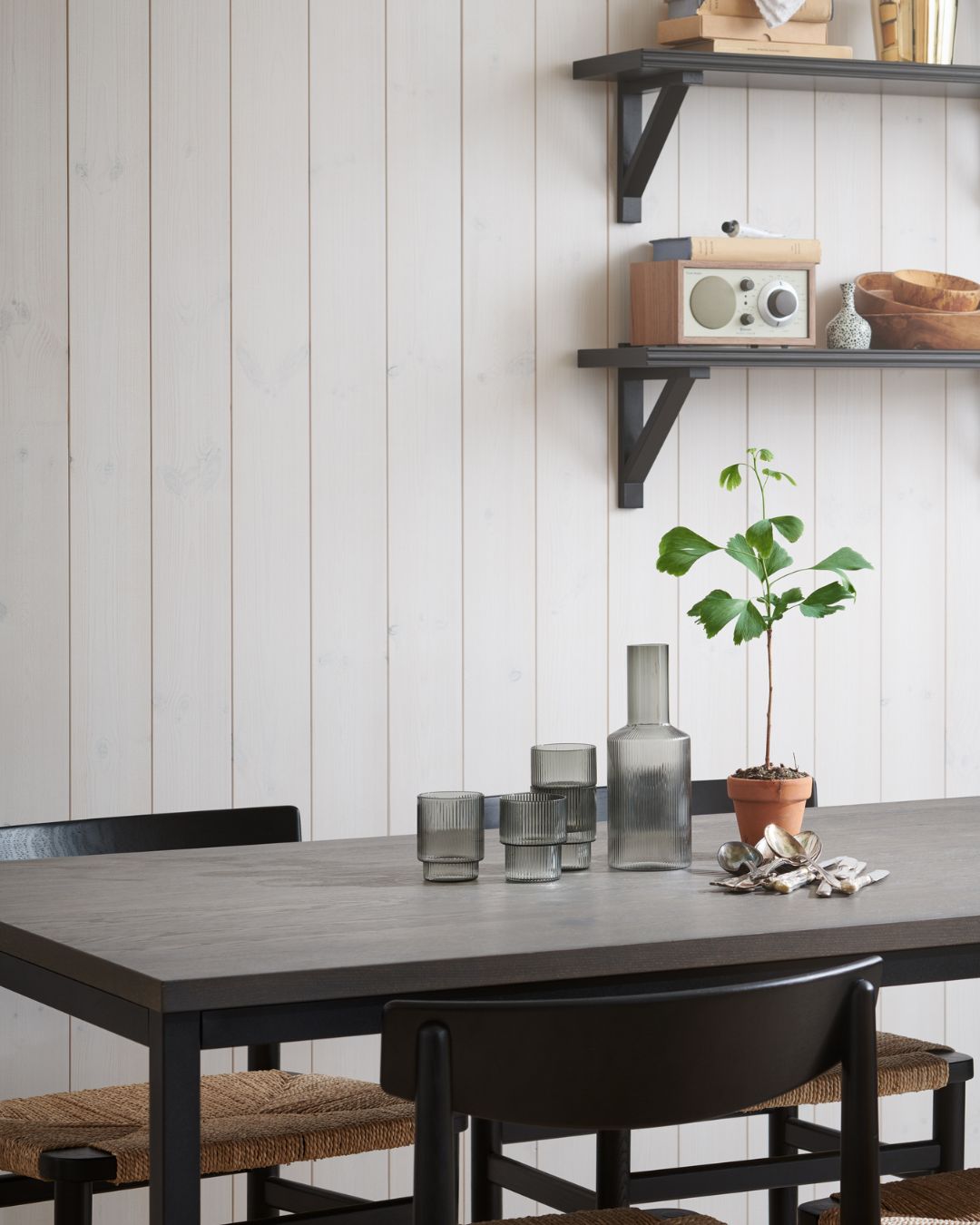 Matplats med vita träpanelen Sjöbod Snö på väggen och mörkt matbord med svarta stolar. På bordet står glasvaser och en liten krukväxt, och ovanför sitter öppna hyllor med böcker, skålar och en radio.