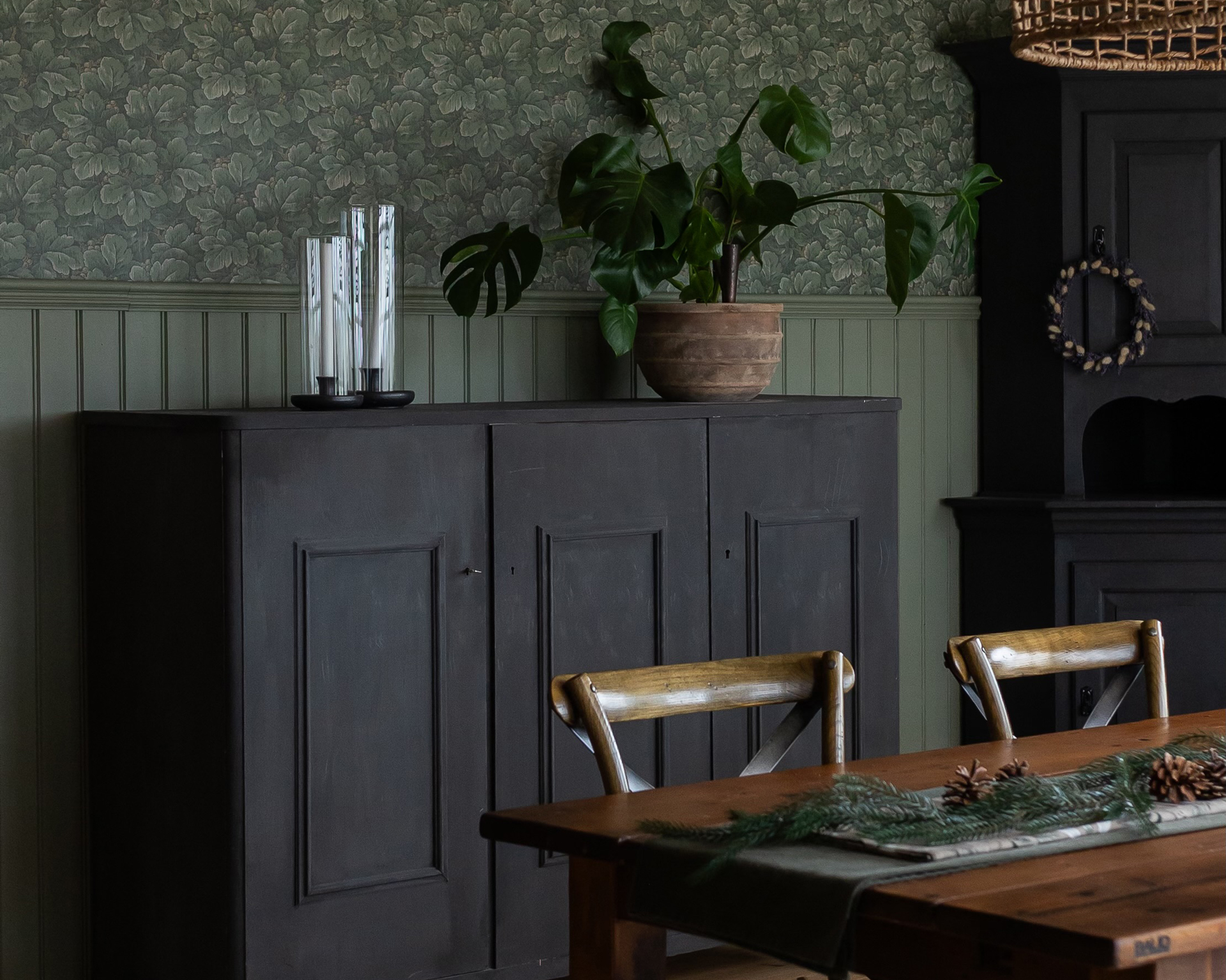 Matsal med vit bröstpanel i pärlspont. Ett brunt matbord och stolar i trä. En fåtölj teddypläd  och ett grått skåp, samt dekorationer