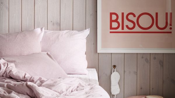Bäddad säng med rosa sängkläder i ett sovrum med grå träpanel och en tavla med texten Bisoui på väggen