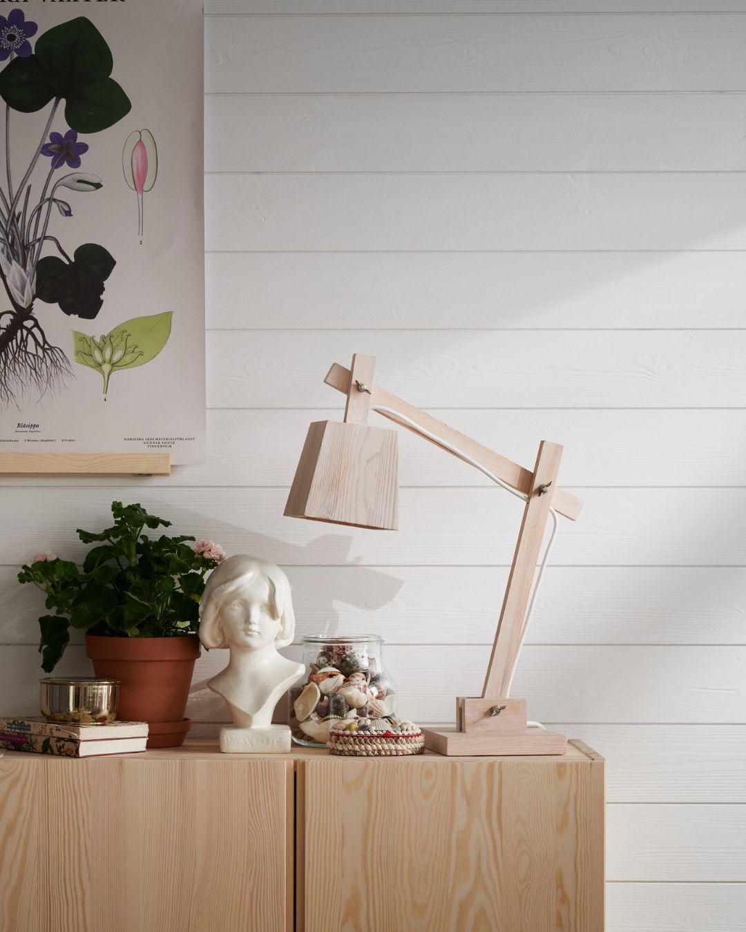 Bänk och bordslampa i ljust trä. En tavla med blommotiv hänger på en vägg med liggande vit träpanel
