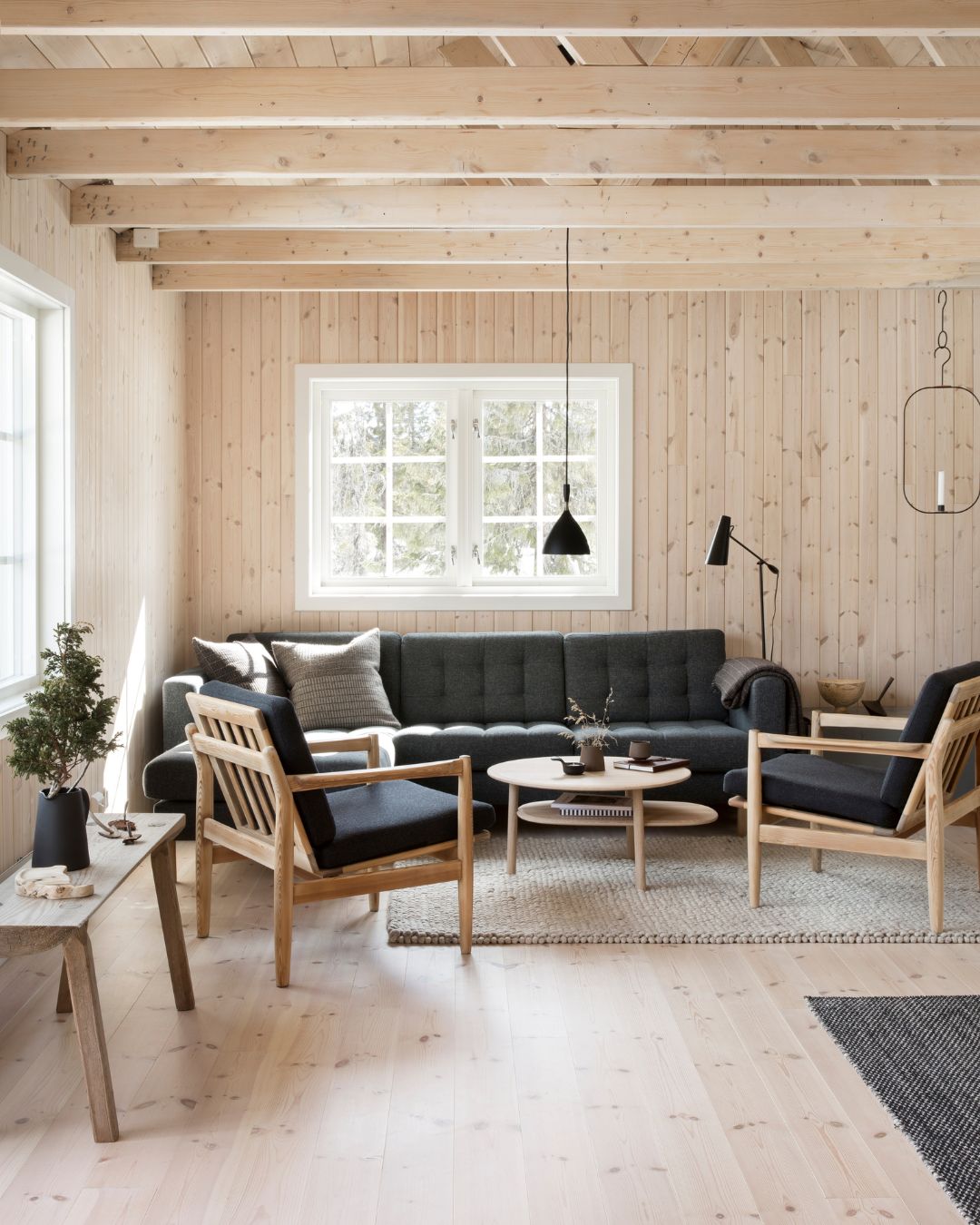 Storstuga i fritidshus med vitvaxad träpanel på väggarna och en mörk soffa framför ett fönster