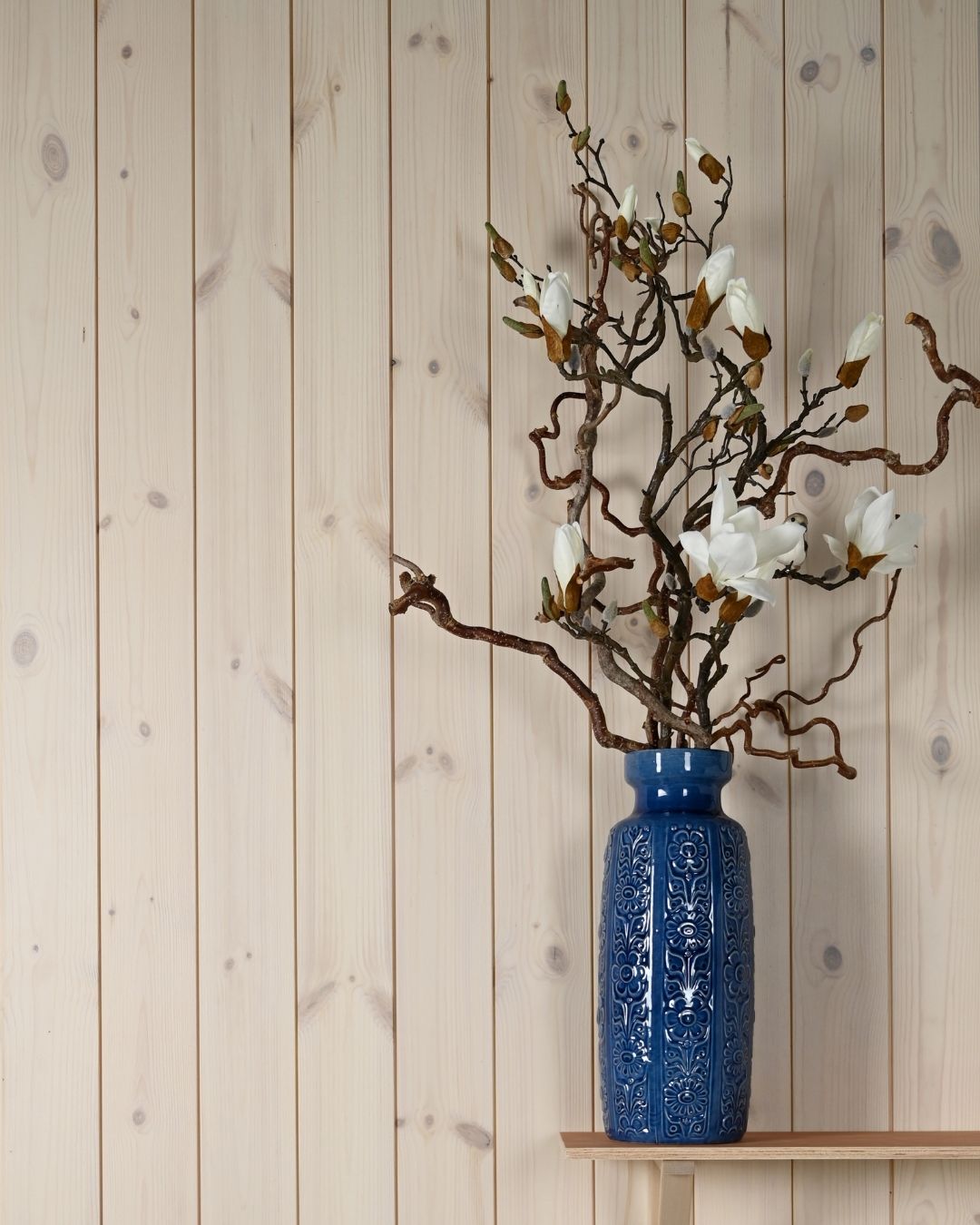 En blå vas med vit magnolia står på en trähylla som hänger på en vägg med stående trävit träpanel