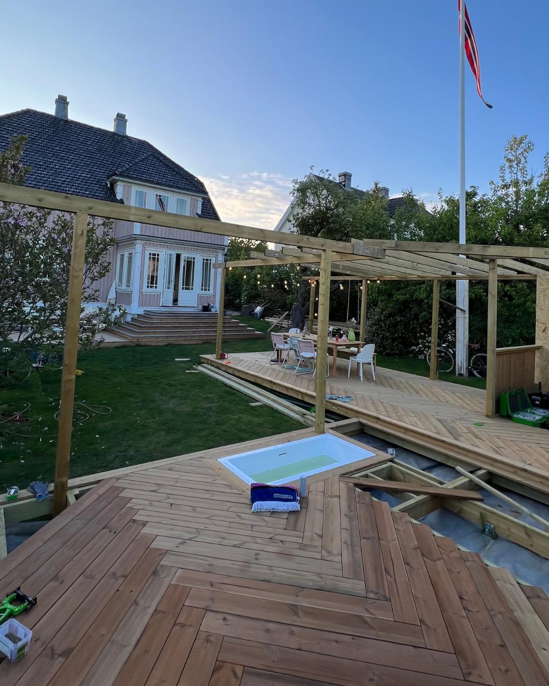 Terrasse under bygging med Moelven Termofuru lagt i fiskebensmønster. Pergolakonstruksjon i bakgrunnen, nedsenkbar bassenglukeåpning synlig i forgrunnen. Rosa trehus og norsk flagg i bakgrunnen.