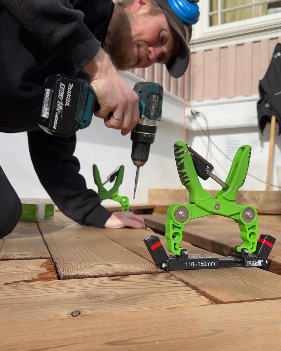 Mann som skrur terrassebord med skrutrekker. Grønne terrasseklemmer av merket ESSVE holder bordavstanden på plass. Nærbilde fra gulvnivå.