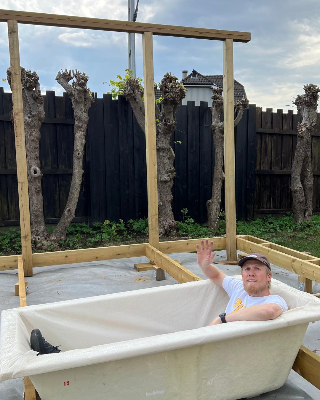 Mann som sitter i et nedsenkbart badekar under installasjon, omgitt av treramme og pergolastolper under bygging. Mørkt tregjerde og klatreplanter i bakgrunnen.