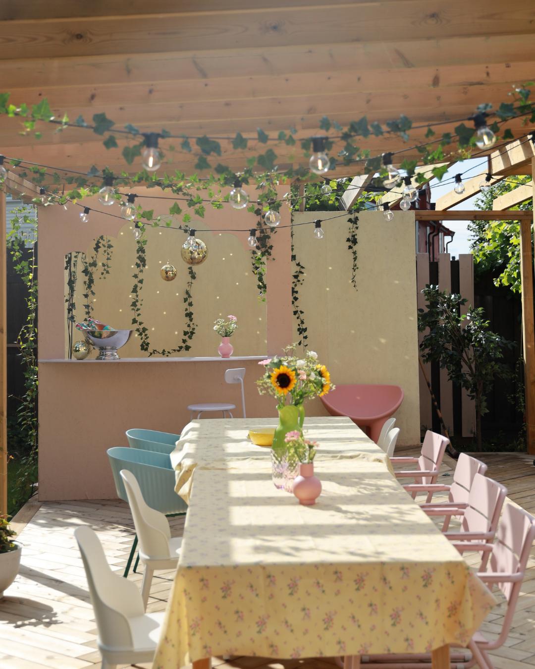 Uteplass med pergola i lyst trevirke, dekorert med lyslenker, eføy og diskokuler. Langt bord med blomstrete duk, solsikker i vase og fargerike stoler i rosa og blått.