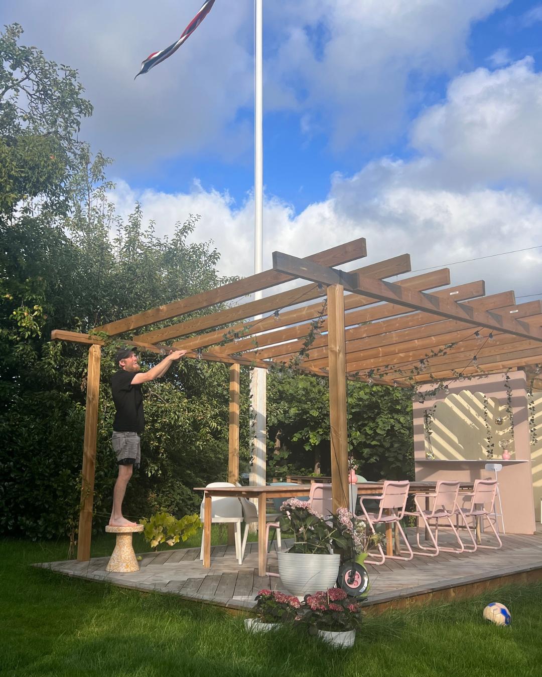 Mann som står på krakk og jobber med lyslenker under en trepergolaen på en terrasse. Norsk flagg vaier i bakgrunnen under blå himmel.