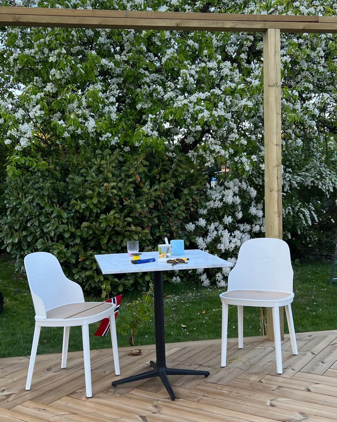 To hvite stoler og et lite bord på en terrasse av Moelven Termofuru. Pergola i bakgrunnen, hvitt blomstrende tre fyller bildet bak.