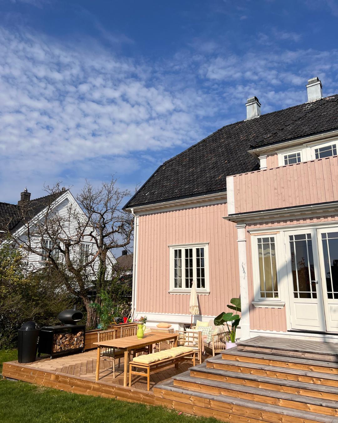 Utsikt mot baksiden av et rosa trehus med terrasse i Moelven Termofuru. Utemøbler i tre og sort grill, blå himmel med lette skyer.