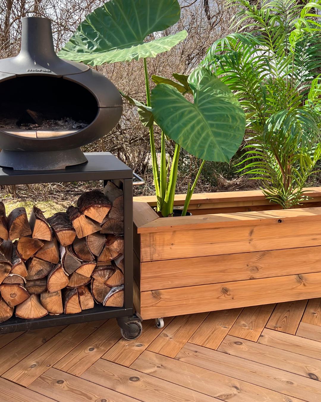 Sort vedovn med vedlager på hjul ved siden av en stor plantekasse i tre, på terrasse av Moelven Termofuru. Tropiske planter i forgrunnen, tidlig vår med bar skog i bakgrunnen.