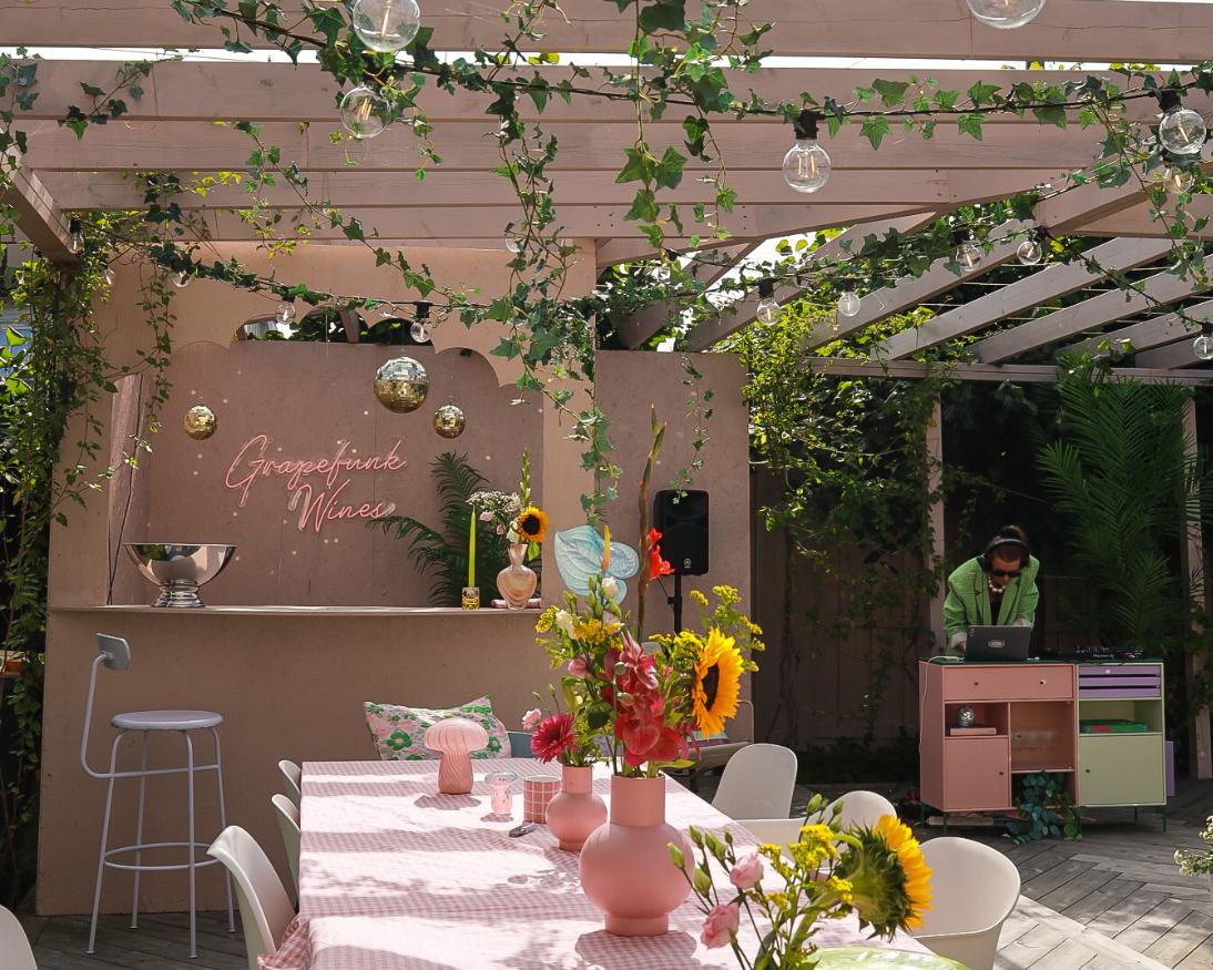 Uteplass under pergola dekorert med lyslenker, eføy og diskokuler. Langbord med rosa rutete duk og solsikker i vaser, bardisk med neonskilt i bakgrunnen og DJ ved miksebord til høyre.