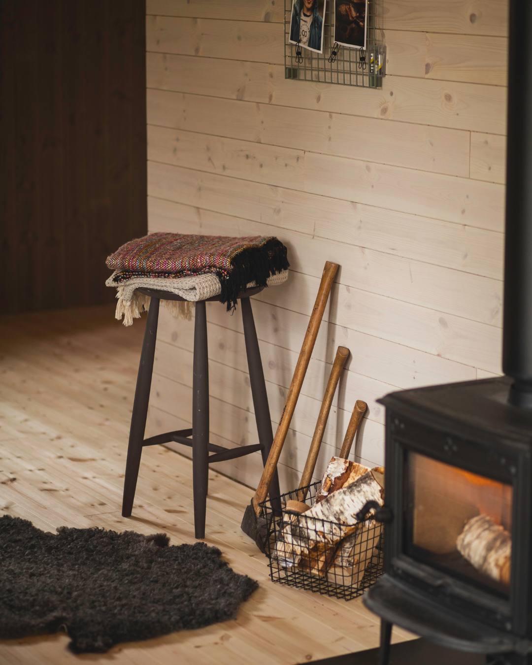 Ett rum med fårskinn på golvet, en hög pall, en kamin och några yxor som lutar mot en vägg med träpanelen Stilla Morgondis