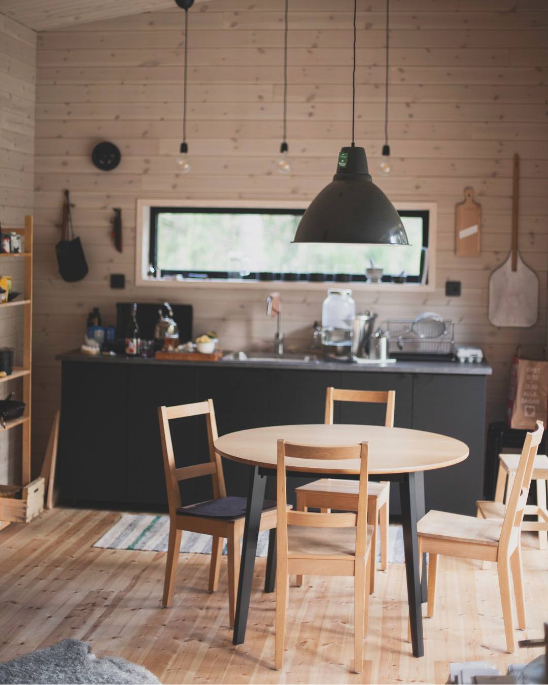 Kjøkken i trivelig svensk stuga med rundt kjøkkenbord i lyst tre, 4 enkle trestoler og en svart lampe. Hytta har lysbeiset panelvegger og furugulv.