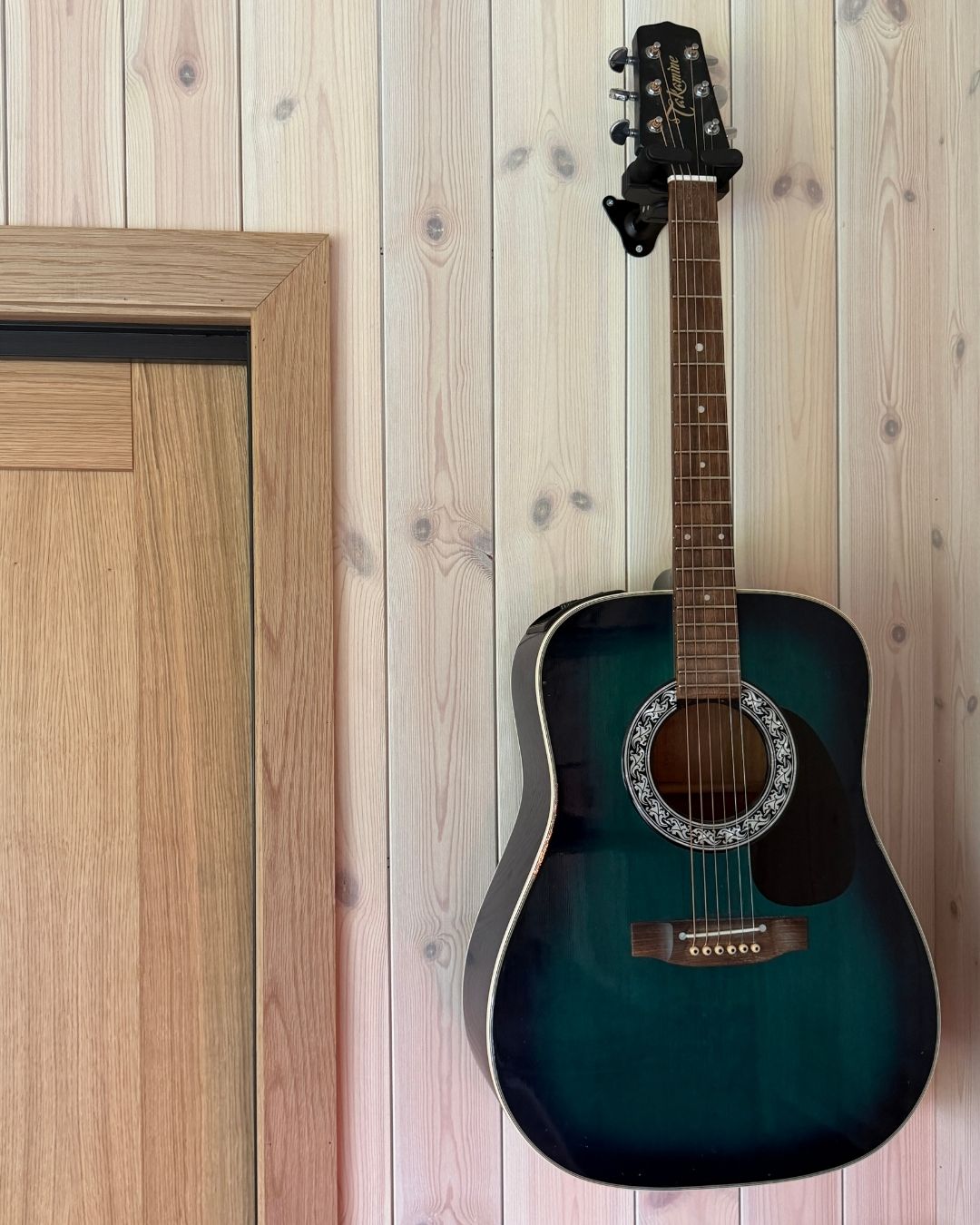 Mørk akustisk gitar henger på en vegg med lyst trepanel ved siden av en tredør.