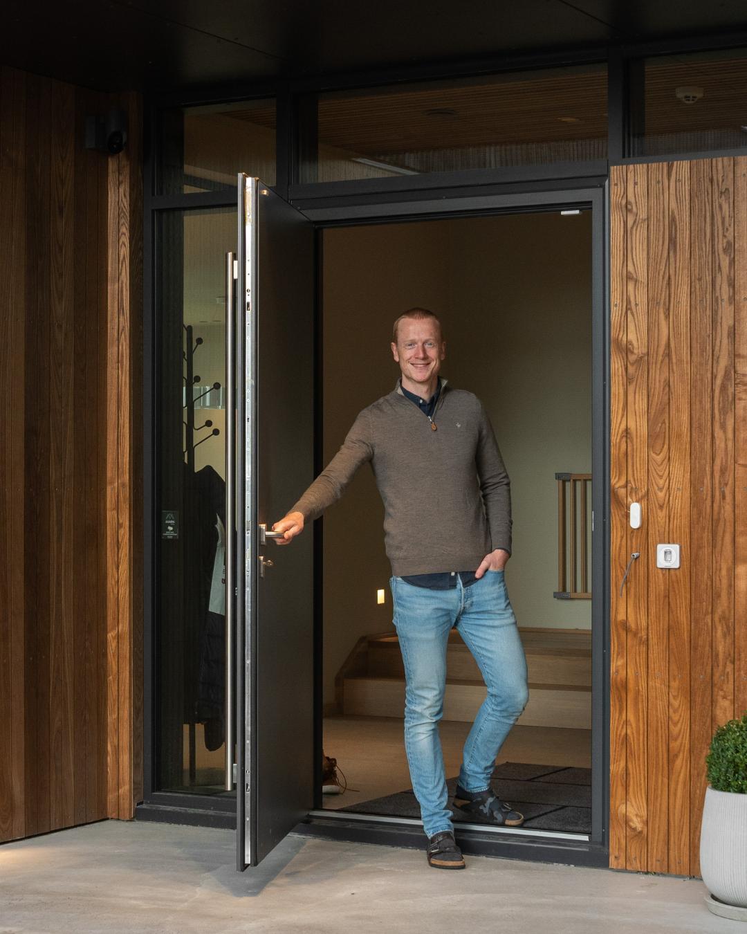 Johannes står smilende i den åpne inngangsdøren til boligen, omgitt av termoask-kledning på veggen og svarte vindusrammer.