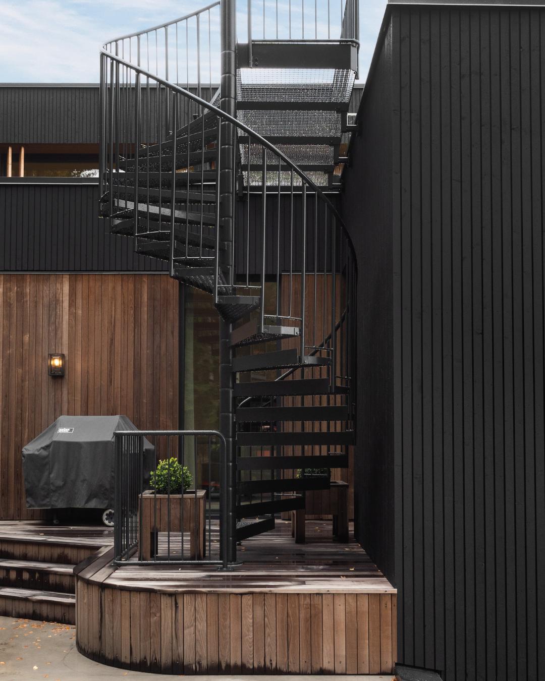 Svart spindeltrapp i metall på utsiden av boligen, flankert av svart Heftig PRO-kledning og termoask-panel. En innebygd platting i tre med trapper og en Weber-grill ses i forgrunnen.