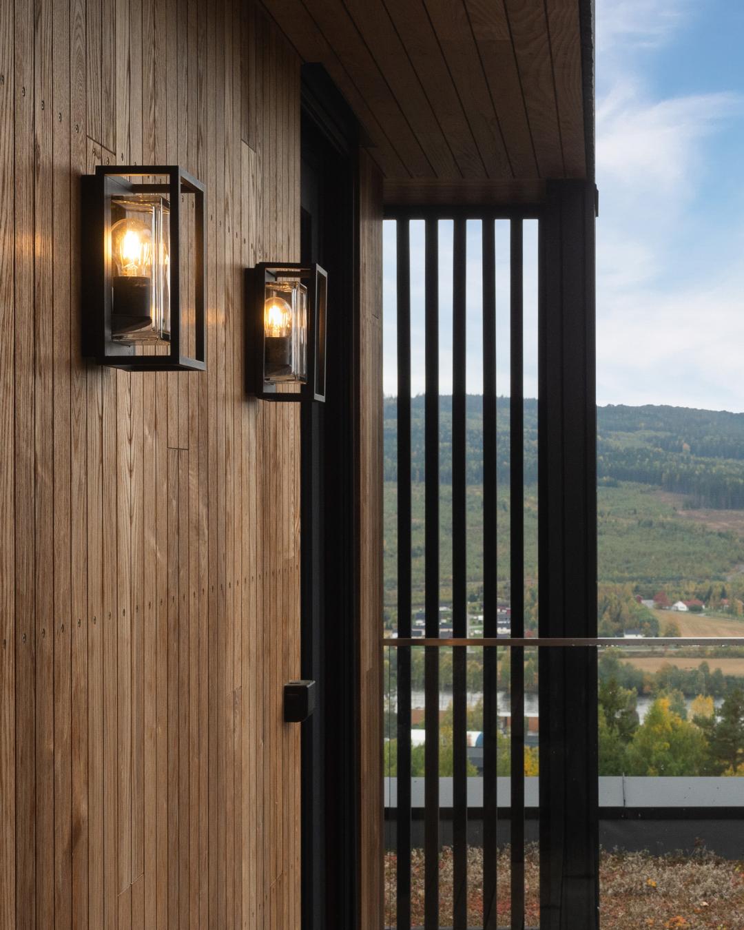 To svarte vegglamper med Edison-pærer montert på stående termoask-kledning ved en overdekket terrasse. Svarte spiler og grønt åslandskap skimtes i bakgrunnen.