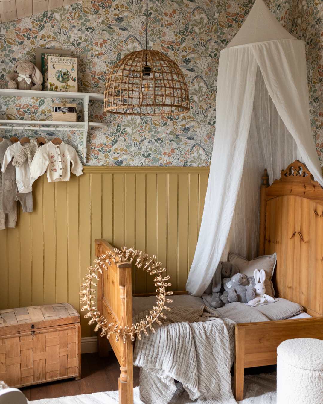 Barnrum med gulmålad pärlspont på nedre delen av väggarna och blommönstrad tapet ovanför. Rummet har en träsäng med vit sänghimmel, en vägghylla med barnkläder och leksaker samt en flätad taklampa i naturmaterial.