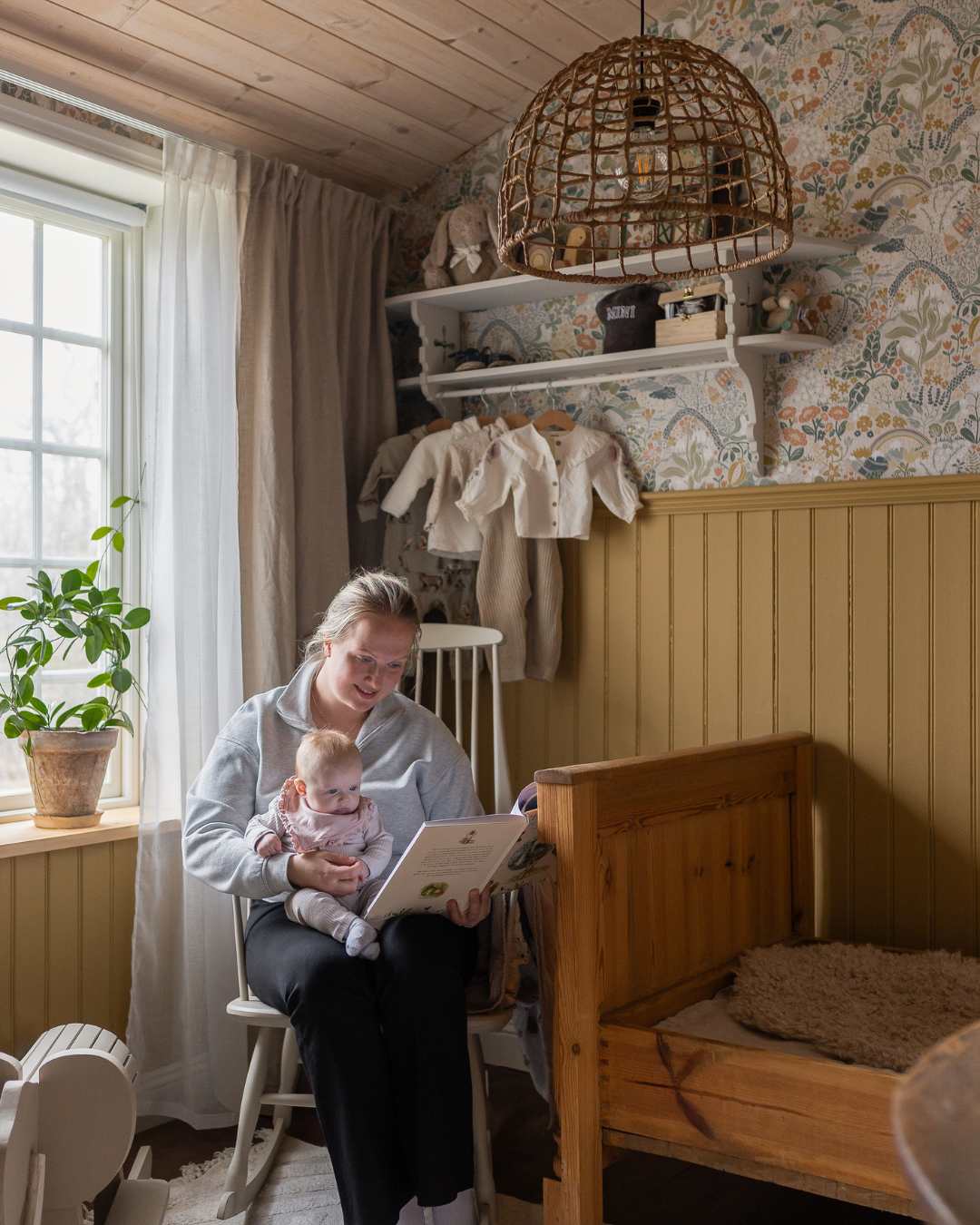 Mor och barn läser bok i barnrum med grön bröstpanel, blommig tapet och träsäng med sänghimmel.