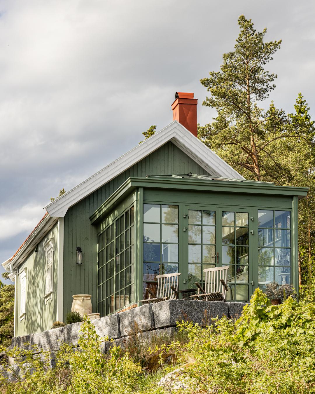 Lite grønnmalt trehus med orangeri og store sprossevinduer, omgitt av naturstein og frodig vegetasjon.