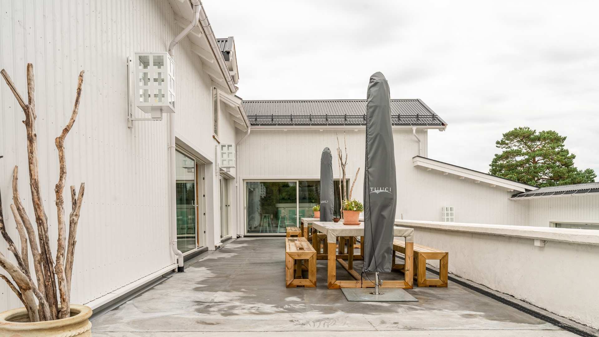 Hvitt hus med stående trekledning og støpt uteplass med parasoll benk og bord. Trepinner i potte brukt som dekor 