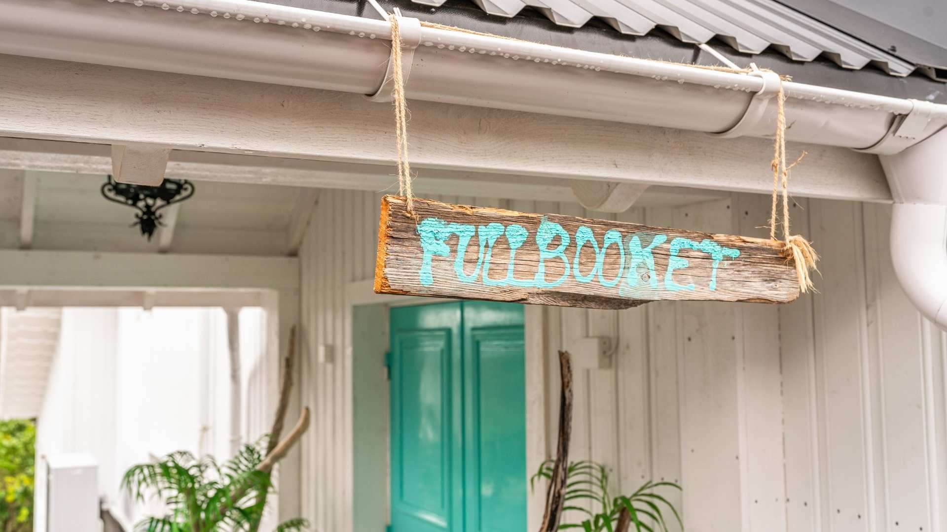 Skilt av drivved  med turkis håndskrift som sier Fullbooket. I bakgrunn er det hvitmalt kledning og en turkis inngangsdør.