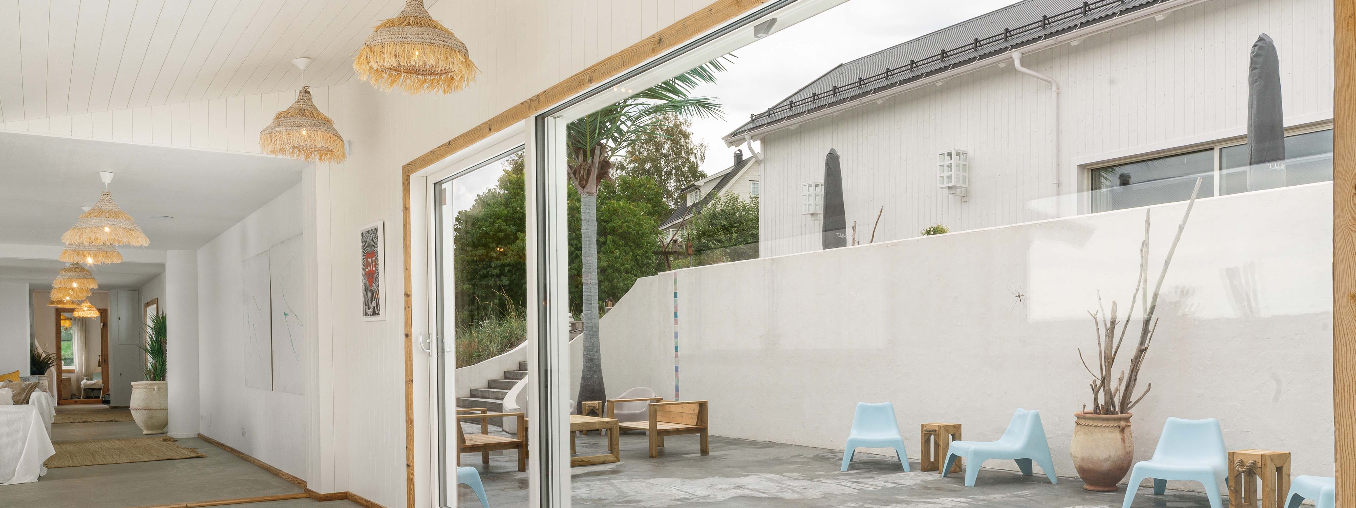 Allrom med hvitmalt sprekkpanel med store åpne terrassedører ut mot gårdsrom. Utvendig  hvitmalt kledning, støpt terrassedekke og fargerike terrassemøbler.