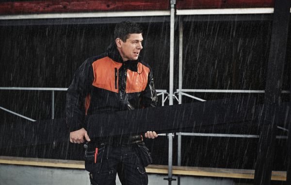 En mann iført arbeidsjakke i sort og oransje håndterer Heftig kledning utendørs i kraftig regnvær. Han holder i en planke og arbeider på et bygg- eller anleggssted, med mørk bakgrunn og synlige stålkonstruksjoner.