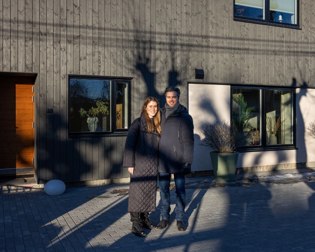 Et par står foran fasaden av Heftig Pro i fargen Labrador en klar vinterdag. Bak dem ses svarte vinduer, hvit murpusset nisje og en naturtre inngangsdør. Treskygger faller dramatisk over fasaden i vinterlyset.