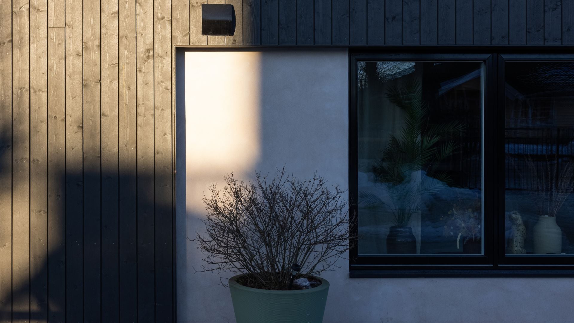 Hushjørne i skumringen med Heftig Pro i fargen Labrador. Veggmontert utelampe kaster varmt lys mot hvit murpusset nisje. Pottet busk foran veggen og svakt opplyst vindusparti.