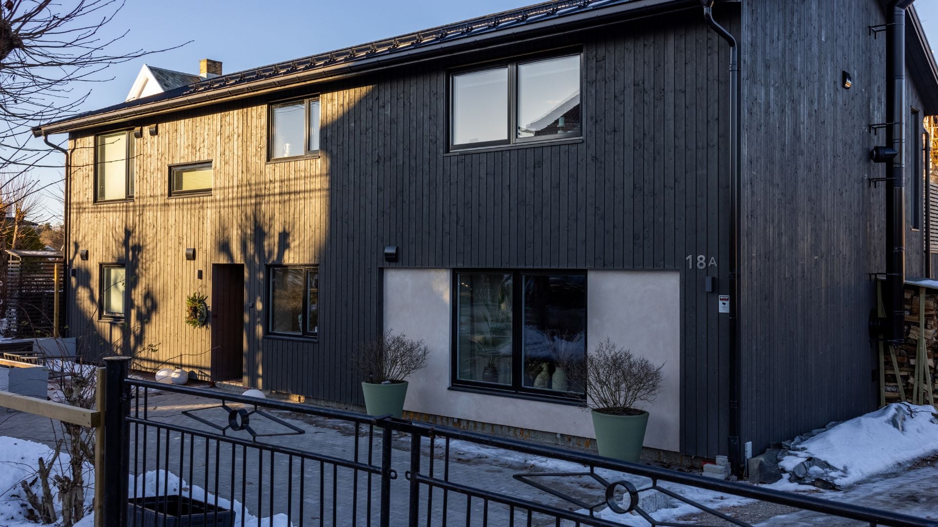Enebolig i to etasjer med fasade av Heftig Pro i fargen Labrador. Store vinduspartier innrammet av hvit murpuss, svarte vinduer og takrenner. Snødekt hage i vinterlys.