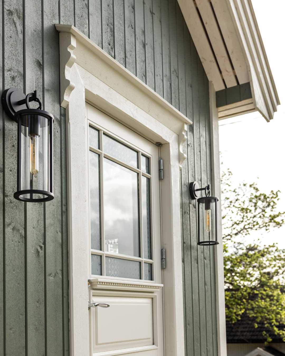 Inngangsparti med stående trekledning i fargen grønn, hvit inngangsdør og hvit omramming på døren. Svarte utelamper