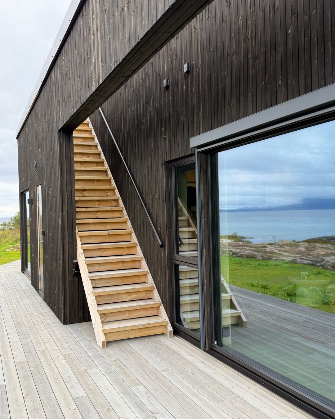 Moderne hus ved sjøen med mørk, stående Værbitt kledning i fargen Garva og lys treterrasse.