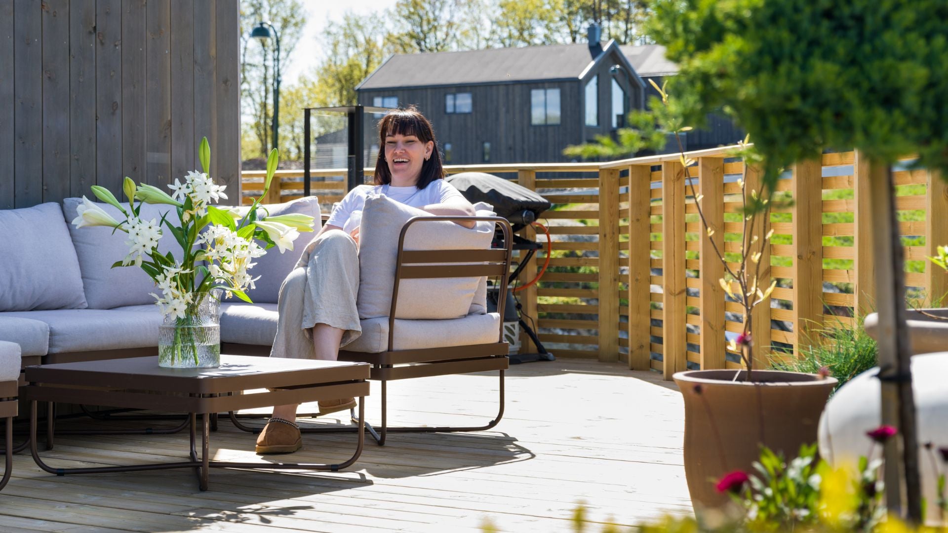 Person sitter i loungestol på treterrasse med sofagruppe, bord og blomster, foran moderne trehus.
