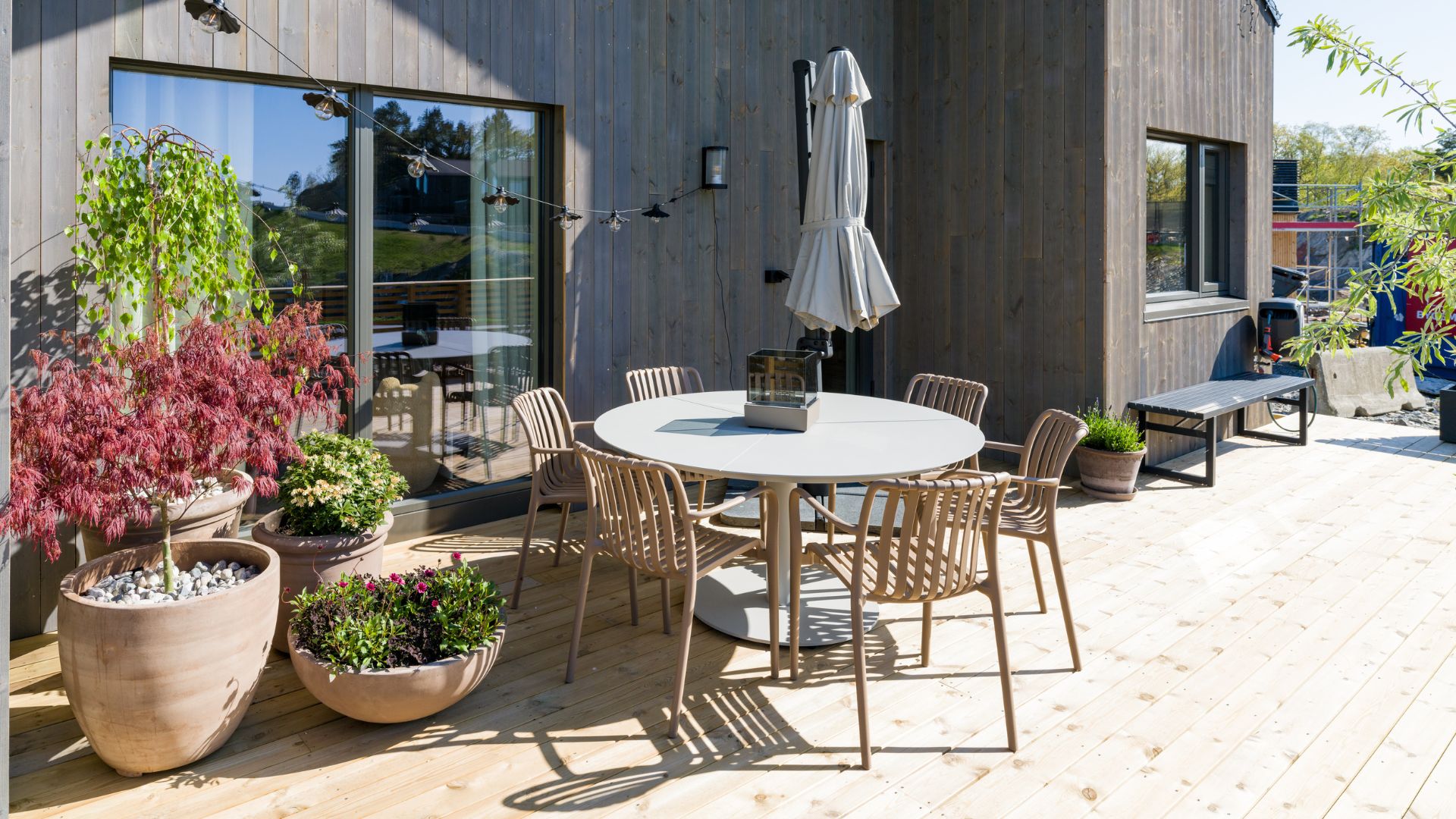Rundt spisebord med flere stoler på romslig terrasse, omgitt av krukker, utemøbler og moderne fasade.