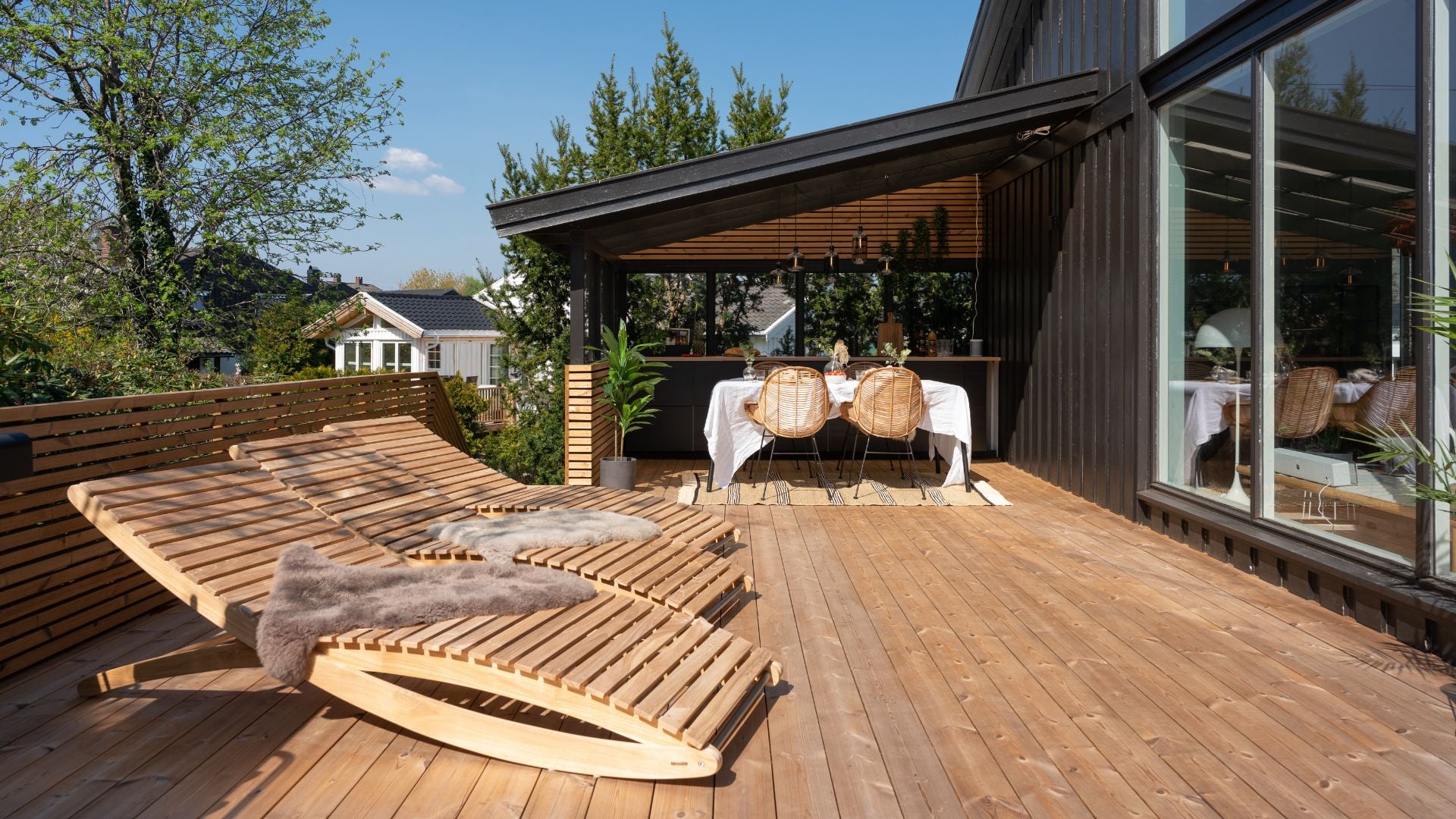 Romslig treterrasse med doble solsenger i tre og spiseplass under tak ved husets fasade.