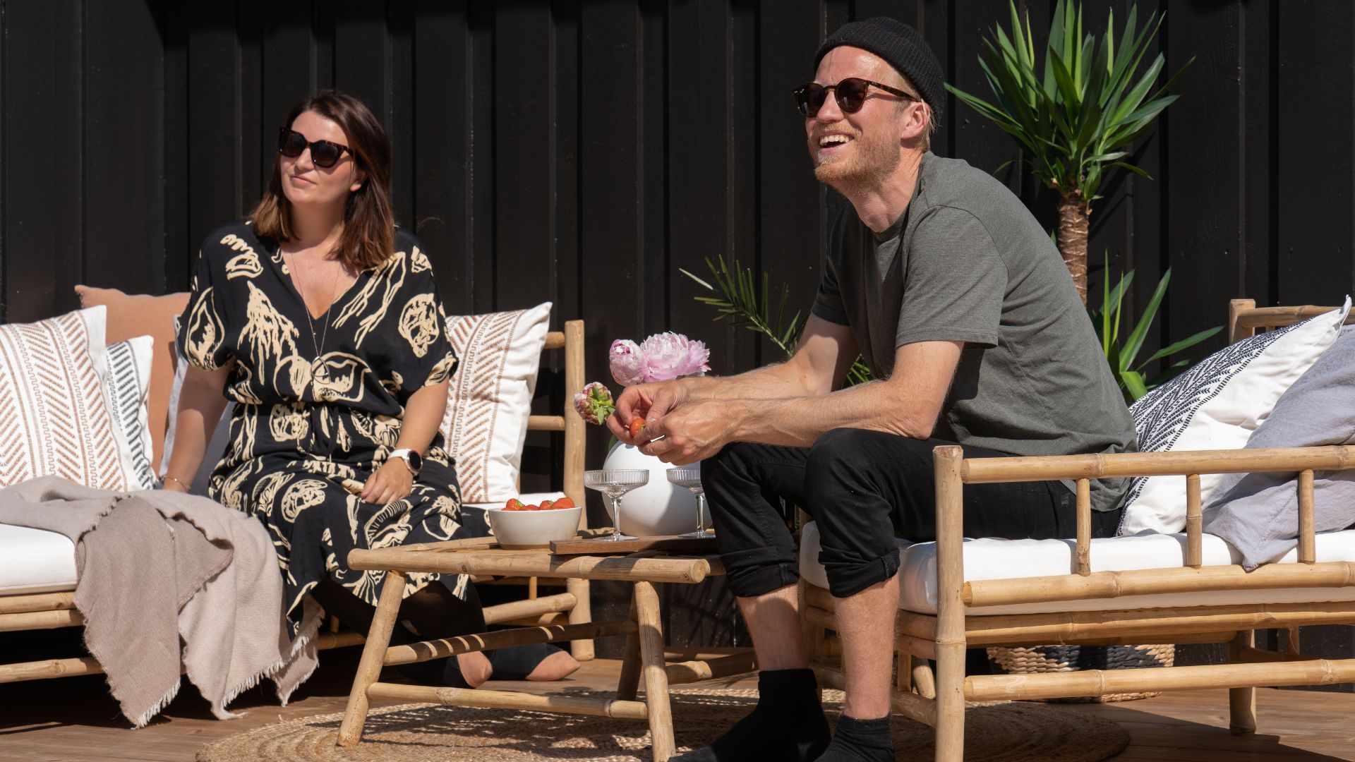 To personer sitter i en loungesofa og lenestol i tre på terrasse, med lavt bord, tekstiler og planter mot mørk vegg.